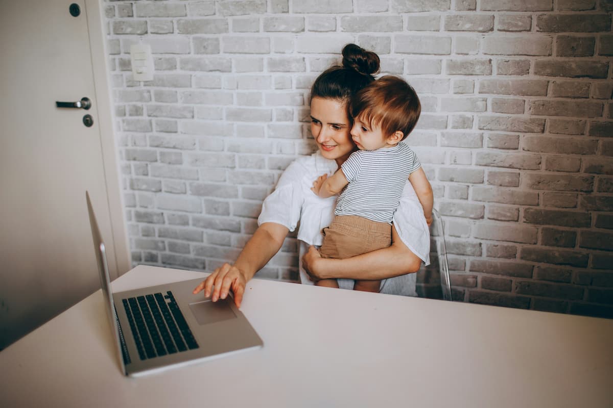 Mãe empreendedora trabalhando no laptop enquanto segura o filho no colo, conciliando rotina profissional e cuidados com a família em casa.