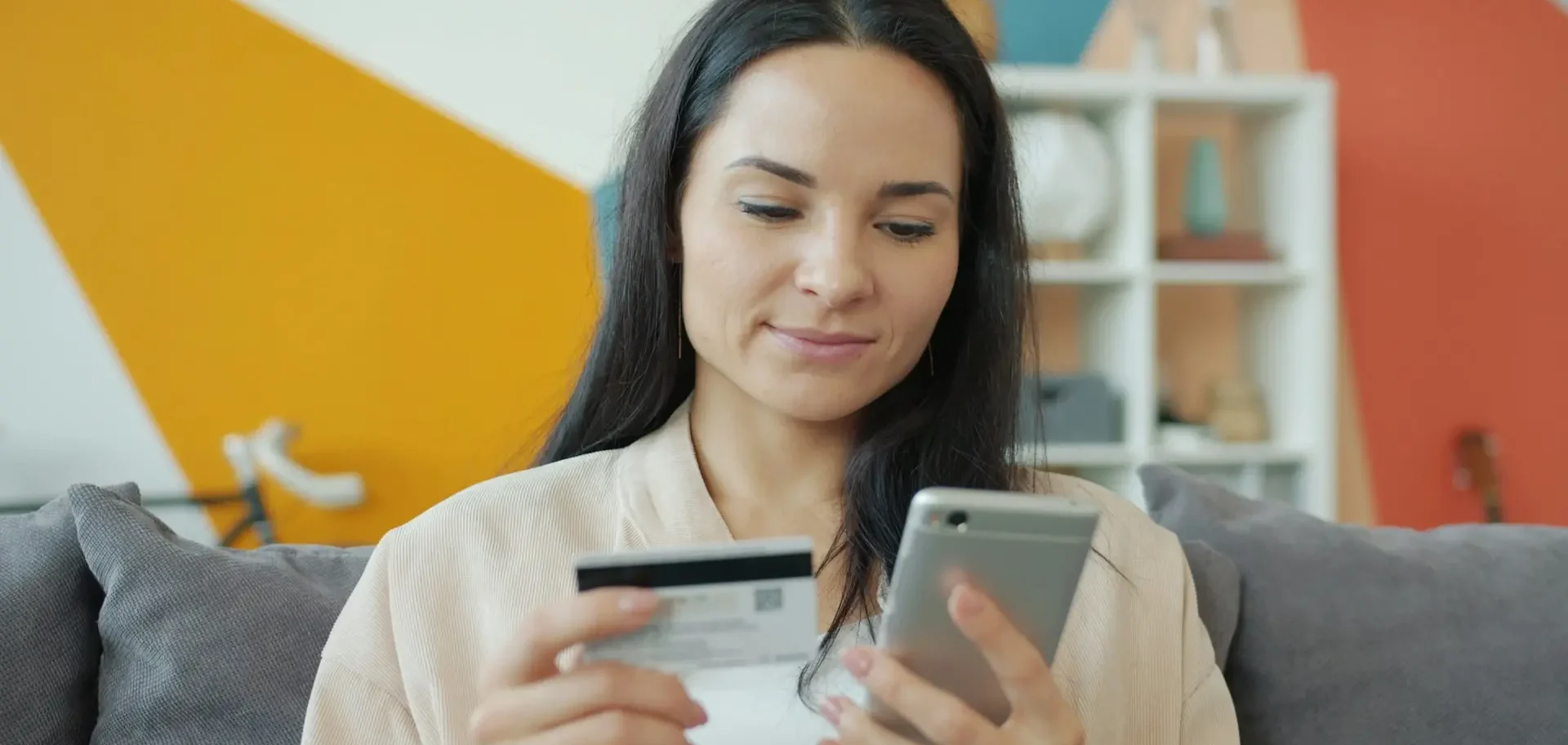 Imagem de uma mulher de cabelos escuros sentada num sofá , segurando um cartão de crédito numa mão e um celular na outra