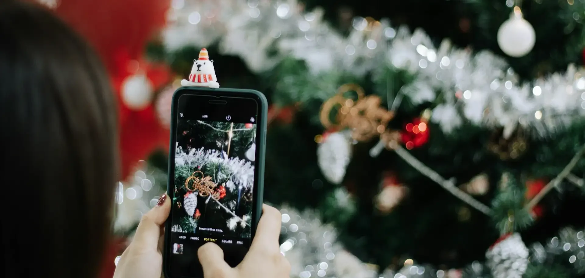 Imagem mostra uma pessoa com o celular na mão tirando uma foto de uma árvore de natal