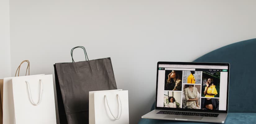 Notebook com página de categoria de produtos de uma loja virtual e sacolas de compras ao lado, representando a navegação em um e-commerce.